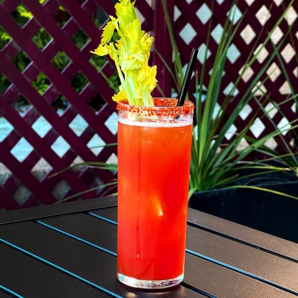 A Bloody Mary cocktail with a celery stalk and chili-salted rim sits on a black table outdoors.