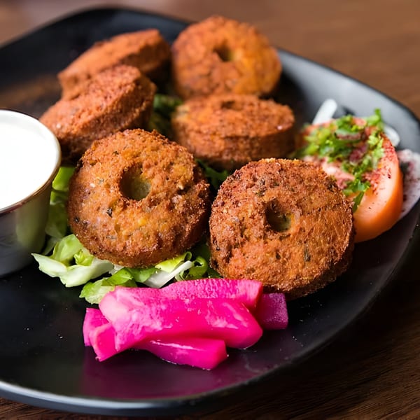 A black plate of Kabob House falafel, with lettuce, tomato, pickled turnips, and a side of white sauce.