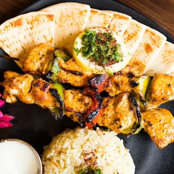 Grilled chicken and vegetable kabobs with rice, pita bread, and a lemon wedge on a black plate.